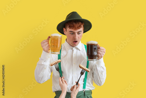 Shocked young man in traditional German clothes holding beer and female hands with sausages on yellow background