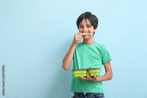 Little boy with lunchbox eating cucumber slice on blue background