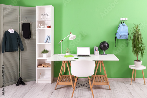 Workplace with desk, shelving unit, laptop computer, backpack and stylish school uniform hanging on folding screen © Pixel-Shot
