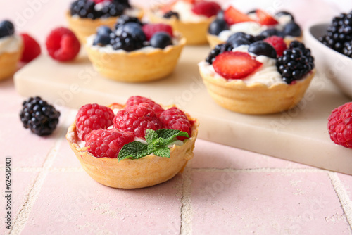 Board of tasty tartlets with whipped cream and berries on pink tile background