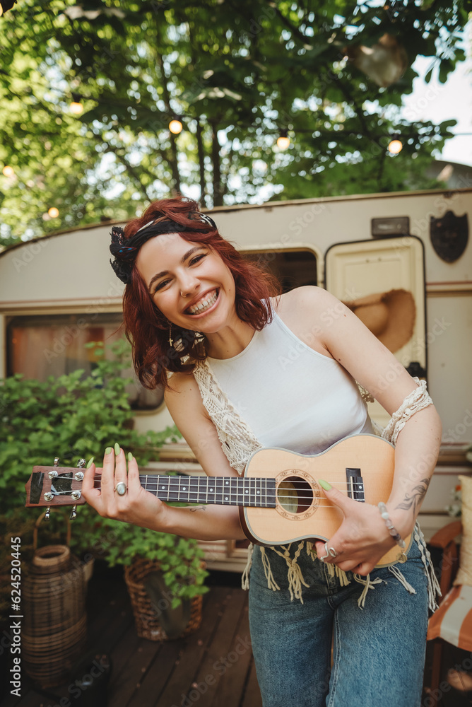 Happy hippie girl is having a good time with playing on guitar in camper trailer. Holiday, vacation, trip concept.