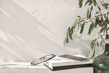 White bookshelf with books, glasses, mobile phone, green vase with plants, empty concrete wall background with aesthetic floral sunlight shadows. Shelf, podium stage mockup, home interior template