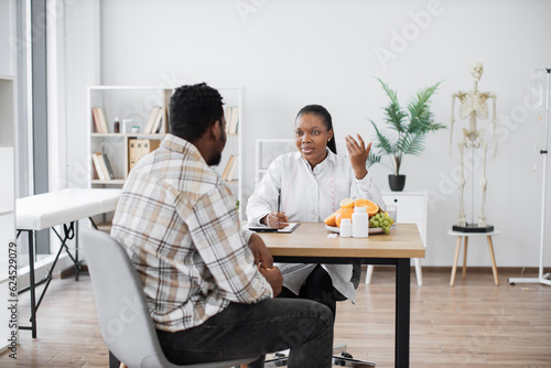 Focused multiethnic woman in doctor's coat talking to middle-aged man in consulting room of hospital. Professional expert in nutrition providing personalized guidance to male client on food habits.
