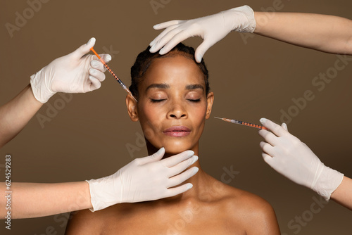 Attractive african american middle aged woman getting facial injections by two cosmetologists, brown background photo