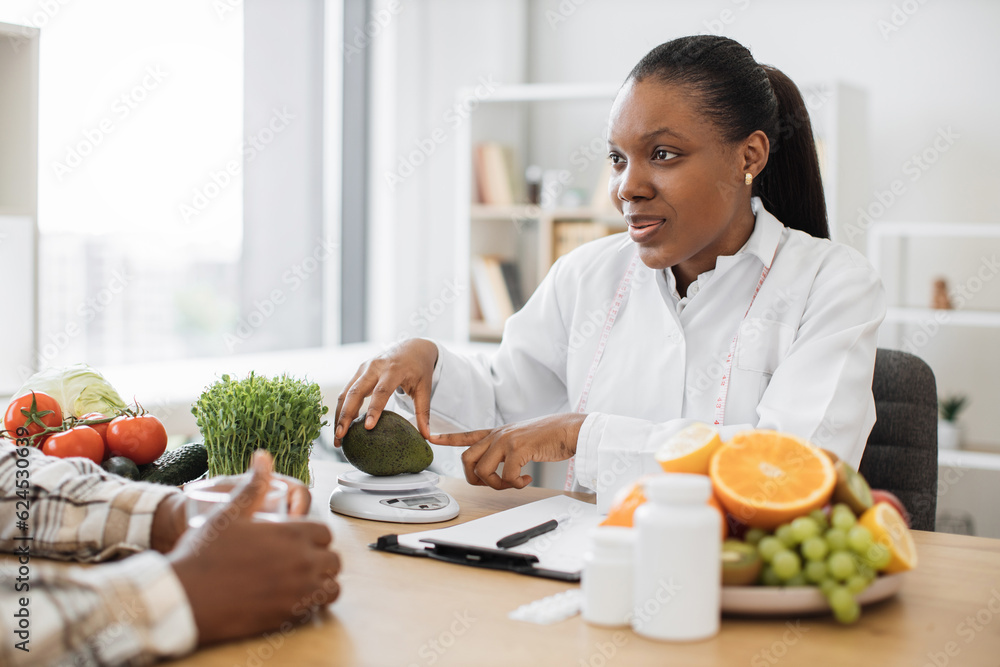 African american dietitian in white coat putting fresh avocado on nutrition scale during patient's appointment. Female professional getting accurate measurements of calories with smart technologies. African american dietitian in white coat putting fresh avocado on nutrition scale during patient's appointment. Female professional getting accurate measurements of calories with smart technologies.