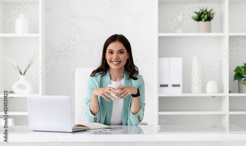 Happy young european woman in suit at table with laptop, enjoys break and cup of coffee