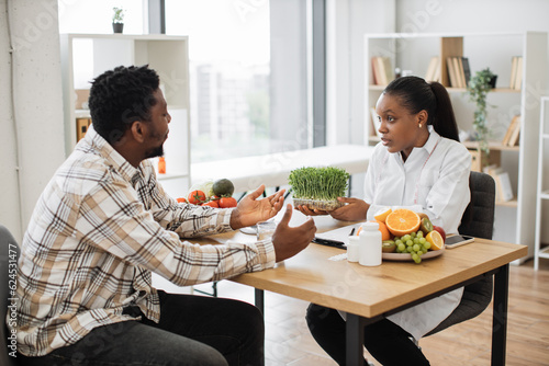 Confident medical specialist showing container with microgreens to adult man in casual clothes sitting at office desk. Multicultural nutrition professional proposing good addition to healthy diet.