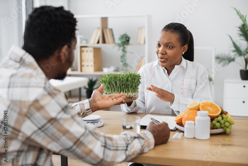 Confident medical specialist showing container with microgreens to adult man in casual clothes sitting at office desk. Multicultural nutrition professional proposing good addition to healthy diet.