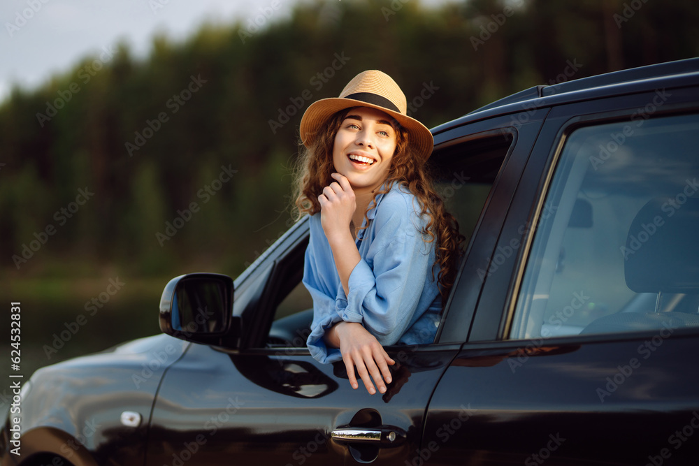 Naklejka premium Happy woman resting and enjoying the sunset leaning out of the car window. Curly tourist woman in a hat has fun and feels freedom on a summer trip. Active lifestyle, nature, tourism.