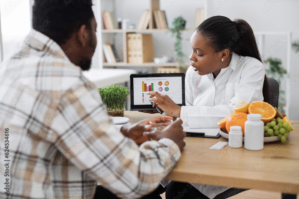 Over-shoulder view of african american woman analyzing patient's nutrition data on digital tablet in consulting room. Efficient dietitian assessing health needs of male client in medical centre. Over-shoulder view of african american woman analyzing patient's nutrition data on digital tablet in consulting room. Efficient dietitian assessing health needs of male client in medical centre.