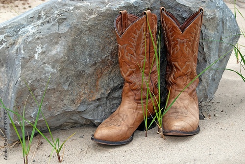 Brown western-style boots on sand by a large rock and green blades of grass 