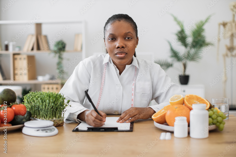 Portrait of african american woman in lab coat holding pen above clipboard while sitting at writing desk in office. Nutrition professional making changes in patient's diet plan in cozy workplace. Portrait of african american woman in lab coat holding pen above clipboard while sitting at writing desk in office. Nutrition professional making changes in patient's diet plan in cozy workplace.