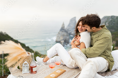 Coastal charms. Loving young couple embracing, drinking wine and enjoying picnic date with beautiful view on ocean shore