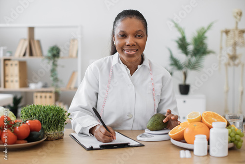 Focused multiethnic female weighing avocado in digital scale while making notes on paper at office desk. Effective dietitian in doctor\'s coat getting accurate measurements using modern device.