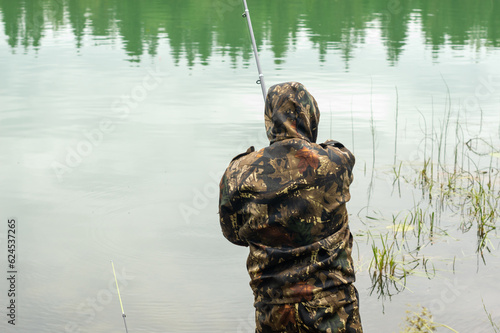 A young male fisherman stands on the shore of the lake with a fishing rod in his hands rear view