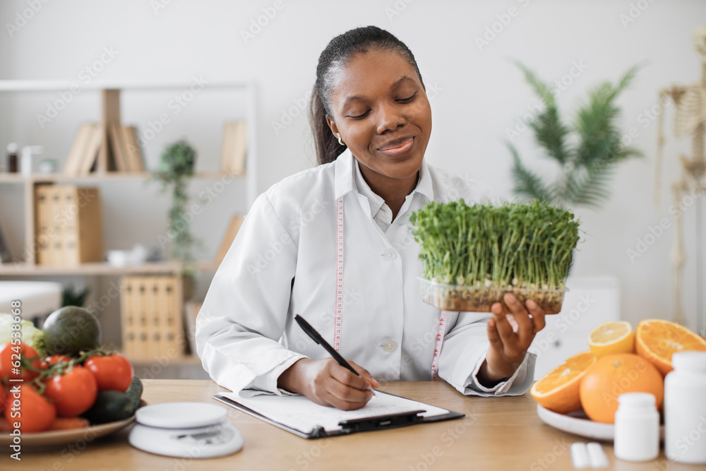 Positive female nutritionist with young sprouts in hand creating meal diet at writing desk in doctor's office. African american specialist with measuring tape recommending clients slimming foods. Positive female nutritionist with young sprouts in hand creating meal diet at writing desk in doctor's office. African american specialist with measuring tape recommending clients slimming foods.