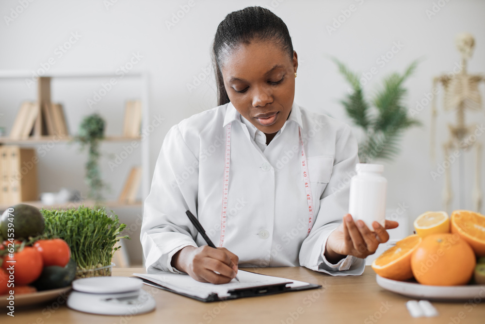 Confident multiethnic female with medicine container in hand recording new ideas of diet plan in workplace. Efficient nutritionist recommending meal modifications with food supplements in hospital. Confident multiethnic female with medicine container in hand recording new ideas of diet plan in workplace. Efficient nutritionist recommending meal modifications with food supplements in hospital.
