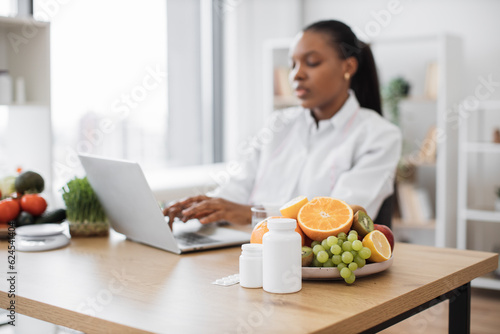 Selective focus of fresh fruits and medications being placed on writing desk at nutritionist s office. Busy african american woman creating educational resources about healthy food choices via device.