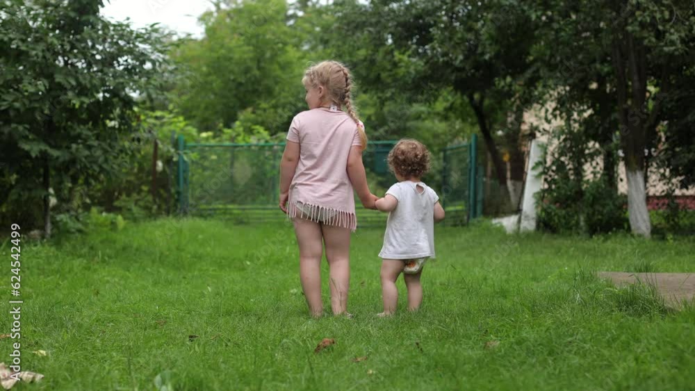 A little girl with bare feet on the grass. Sandals in summer, rest. Green grass, for children, lawn near the house. Little children walk barefoot on the grass. Two sisters holding hands A little girl with bare feet on the grass. Sandals in summer, rest. Green grass, for children, lawn near the house. Little children walk barefoot on the grass. Two sisters holding hands