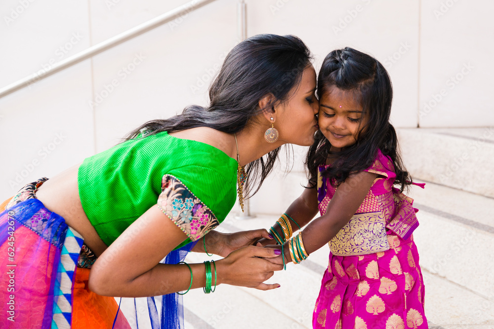beautiful indian family mom mother with daughter girl holding hands kissing and smiling with a bindi and traditional sari dress in front of a building with staircase beautiful indian family mom mother with daughter girl holding hands kissing and smiling with a bindi and traditional sari dress in front of a building with staircase