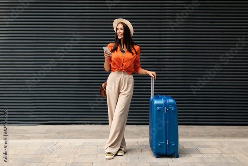Fotografia Cheerful Passenger Woman With Travel Suitcase Using Smartphone Standing Outdoors