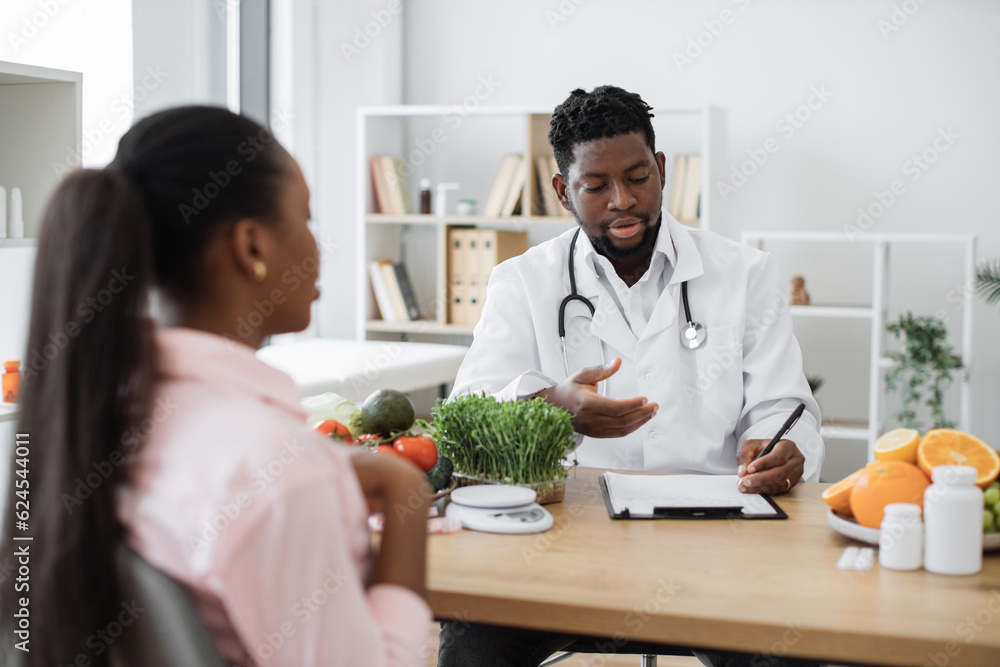 Selective focus of multiethnic man in lab coat sitting at desk with woman in consulting room of hospital. Experienced specialist in nutrition giving recommendations about healthy food choices. Selective focus of multiethnic man in lab coat sitting at desk with woman in consulting room of hospital. Experienced specialist in nutrition giving recommendations about healthy food choices.