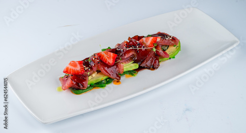 Cold appetizer carpaccio with tuna, avocado, algae, strawberries, spinach, sesame seeds and sauce in a plate.