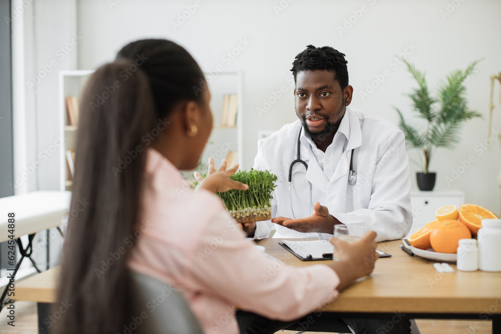 Over shoulder view of african american man with stethoscope proposing micro herbs to pretty woman at work. Medical specialist in food offering young vegetable sprouts as health-supporting component. Over shoulder view of african american man with stethoscope proposing micro herbs to pretty woman at work. Medical specialist in food offering young vegetable sprouts as health-supporting component.