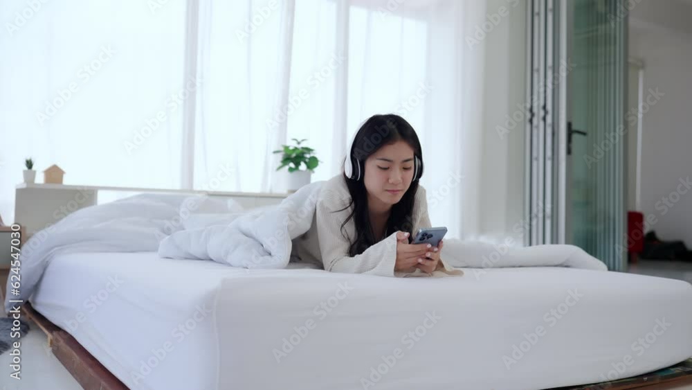Cute Asian woman wearing headphones lying on white bed playing mobile phone.