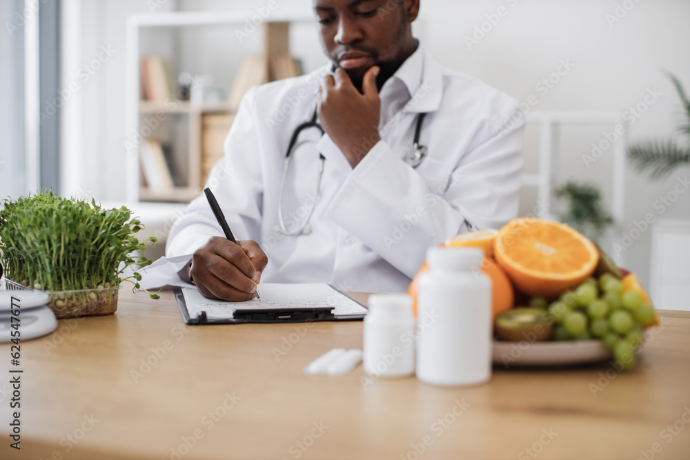Expert in nutrition making special menu for patient and writing list of allowed products on clipboard paper during work time in hospital. African american man touching chin with hand and thinking. Expert in nutrition making special menu for patient and writing list of allowed products on clipboard paper during work time in hospital. African american man touching chin with hand and thinking.