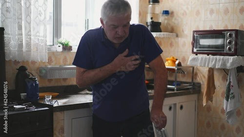 Sickly senior man experiencing a heart attack with chest palpitations at home by kitchen sink. Medically ill elderly person needing emergency aid