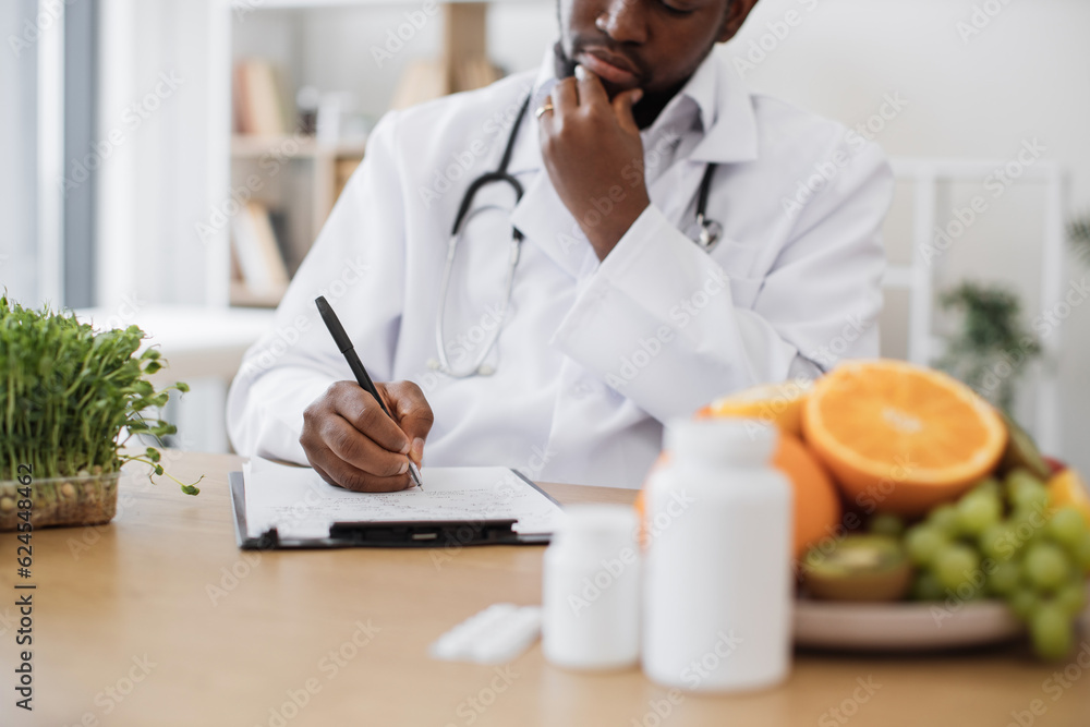 Expert in nutrition making special menu for patient and writing list of allowed products on clipboard paper during work time in hospital. African american man touching chin with hand and thinking. Expert in nutrition making special menu for patient and writing list of allowed products on clipboard paper during work time in hospital. African american man touching chin with hand and thinking.