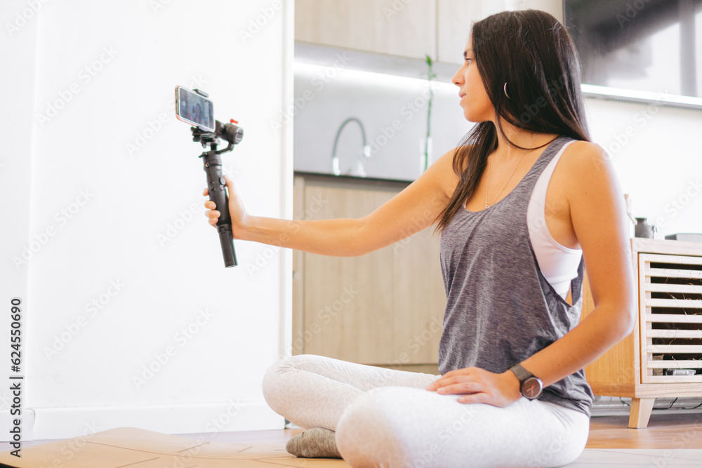 Ταπετσαρία Young Latin woman doing a yoga stream with her mobile phone and a digital stabilizer at home for her networks
