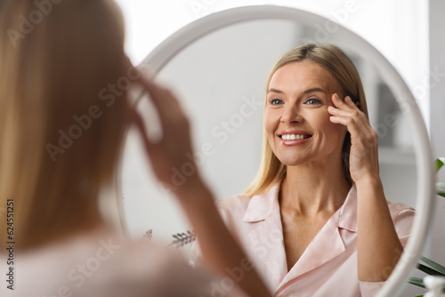 Foto Cheerful mature woman looking at mirror at home, applying eye cream