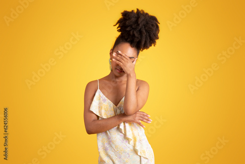 Sad confused unhappy young black woman closes eyes with hand, isolated on yellow background