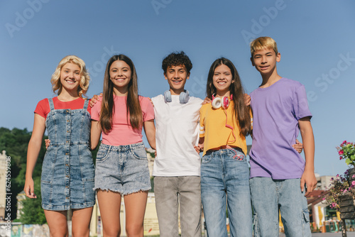 Group of smiling friends, multiracial teenagers wearing colorful t shirts hugging standing outdoors. Happy stylish boys and girls on street looking at camera. Friendship, positive lifestyle, summer