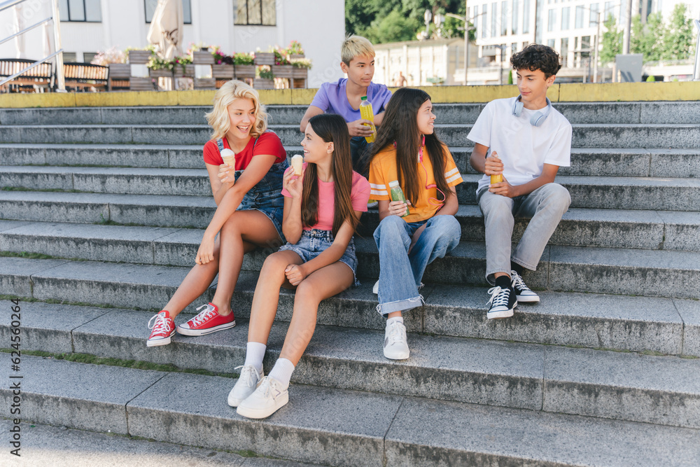 Fototapeta premium Group of smiling friends, multiracial teenagers eating ice cream, drinking lemonade sitting on stairs. Happy stylish boys and girls communication on the street. Friendship, positive lifestyle, summer