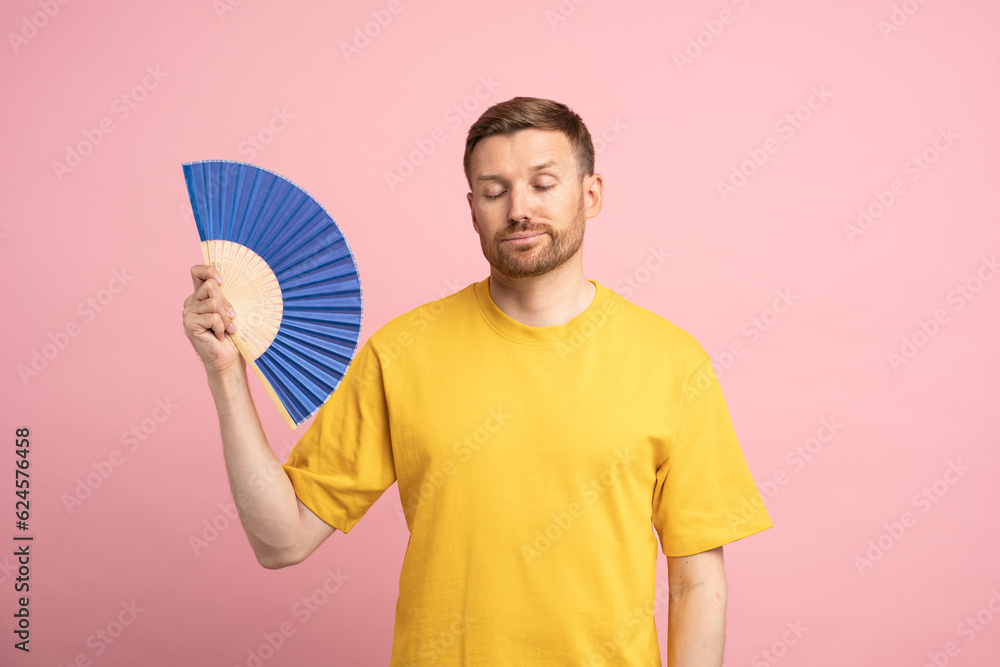 Overheated man waving paper fan suffer from heat, feels sluggish and drowsiness. Unhappy exhausted guy cooling in hot summer weather isolated on studio pink background. Overheating, stuffiness concept Overheated man waving paper fan suffer from heat, feels sluggish and drowsiness. Unhappy exhausted guy cooling in hot summer weather isolated on studio pink background. Overheating, stuffiness concept