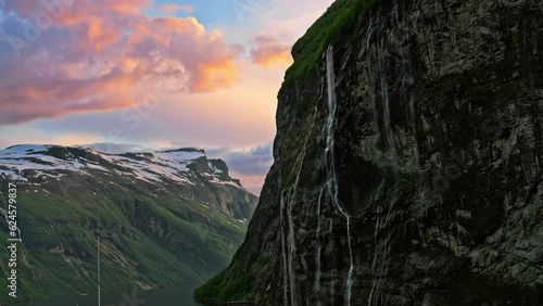 Fantastic picturesque silhouette view of steep snow-capped mountain cliffs with gorgeous waterfalls and dramatic colorful sunset sky on the Geiranger Fjord, Norway