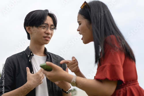 A young asian woman confronts her cheating boyfriend over a extramarital relationship with another girl on his phone. A man taken aback and surprised after getting caught red-handed.