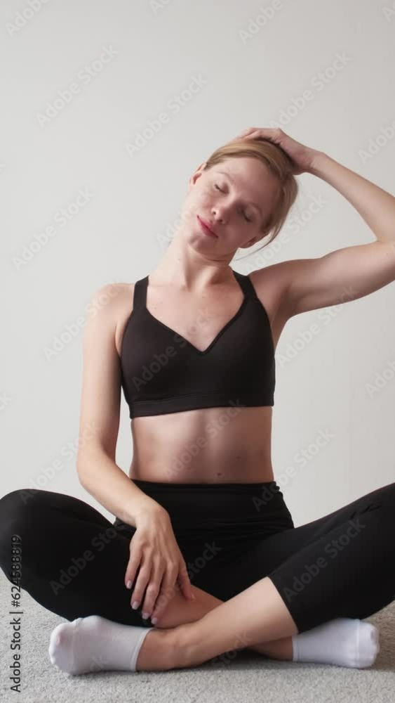 Vertical video. Home yoga. Morning sport. Flexibility training. Relaxed athletic woman stretching neck sitting cross-legged on light free space background. Vertical video. Home yoga. Morning sport. Flexibility training. Relaxed athletic woman stretching neck sitting cross-legged on light free space background.