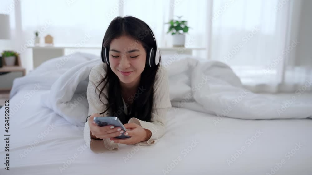 Cute Asian woman wearing headphones lying on white bed playing mobile phone. Cute Asian woman wearing headphones lying on white bed playing mobile phone.