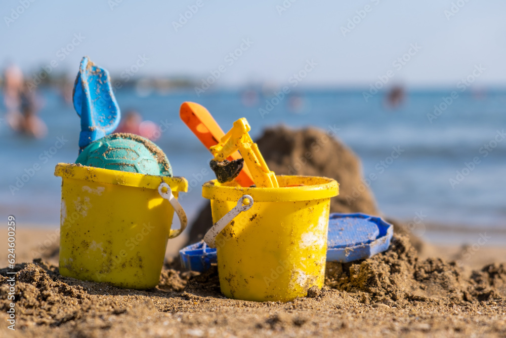 Children's beach toys - buckets, spade and shovel on sand on a sunny day. Bright toys on the background of the sea. Children's beach toys - buckets, spade and shovel on sand on a sunny day. Bright toys on the background of the sea.