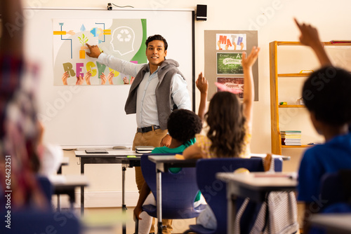 Diverse male teacher teaching and elementary schoolchildren raising hands in class