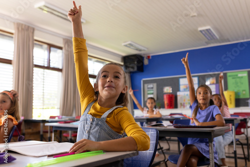 Happy diverse schoolgirls sitting at desks and raising hands in classroom