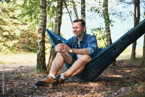 Smiling man messaging on phone while sitting in hammock in forest during summer vacation photo