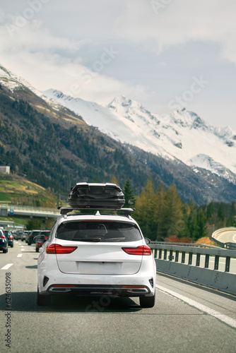 White wagon with roof box storage on the highway in Switzerland. Modern family car adventures in the Alpine Mountains in Europe. Plastic luggage compartment on a car roof. Road trip getaway concept.