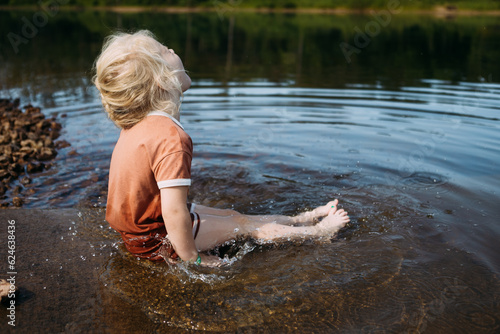 Child sitting in water in casual clothes at edge of pond splashi photo
