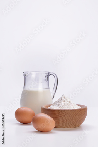 glass jug full of fresh milk with wooden bowl full of white flour and eggs on rustic wooden table