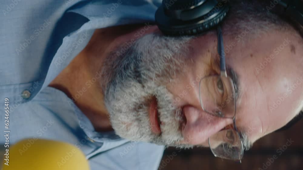 Senior blogger with gray beard in headphones sitting by microphone and speaking while hosting podcast in recording studio. Close-up shot, vertical clip Senior blogger with gray beard in headphones sitting by microphone and speaking while hosting podcast in recording studio. Close-up shot, vertical clip
